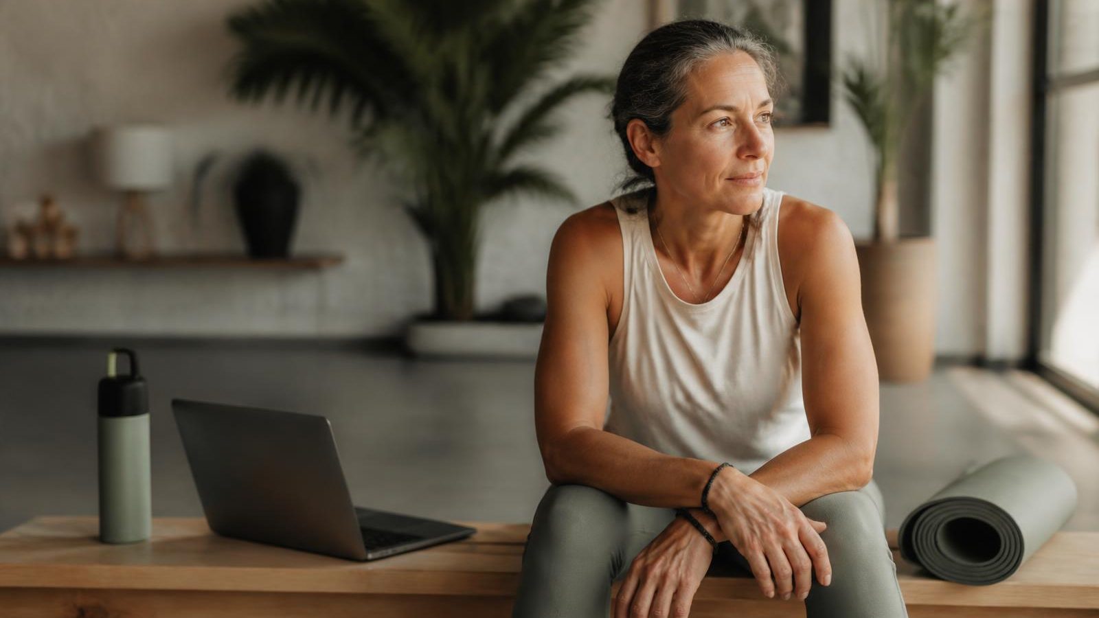 An experienced fitness and wellness professional in her fifties sitting on a bench in a sunlit studio beside an open laptop, a rolled yoga mat and a water bottle, looking thoughtfully toward the window.